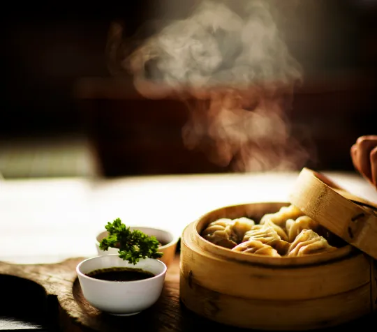Steaming dumplings in a bamboo basket with a hand lifting the lid, accompanied by dipping sauce and garnish in small bowls.