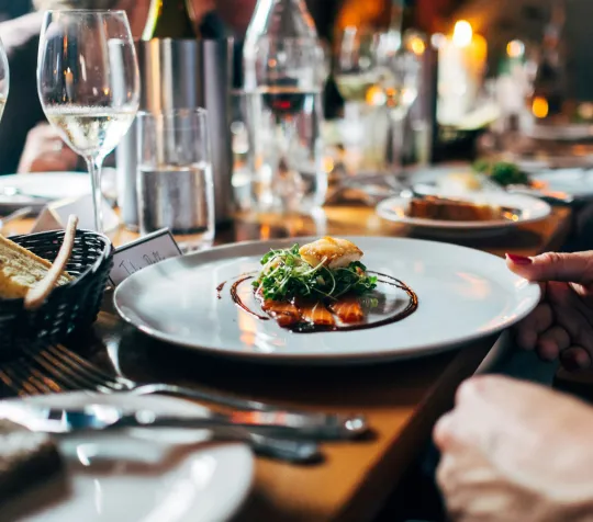 Elegant dining scene with a gourmet dish, bread basket, champagne glasses, and a hand holding a plate on a wooden table.