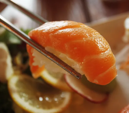 Chopsticks holding a piece of salmon nigiri sushi, with sliced lemon and vegetables blurred in the background.